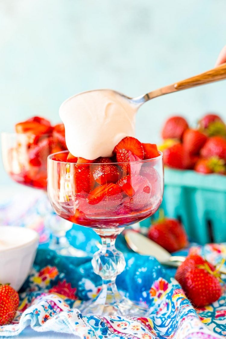 Cream sauce being spoons over macerated strawberries in a stemmed glass to make Strawberries Romanoff.