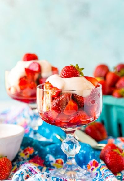 Straight on shot of a stemmed dessert dish with strawberries romanoff in it with more strawberries in the background.
