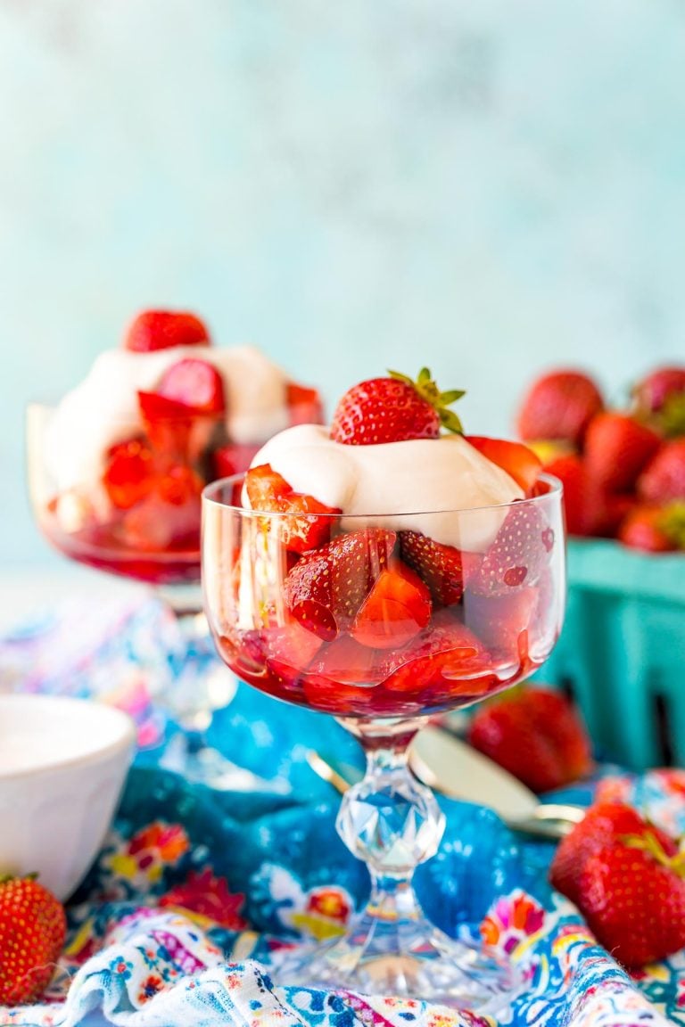 Straight on shot of a stemmed dessert dish with strawberries romanoff in it with more strawberries in the background.