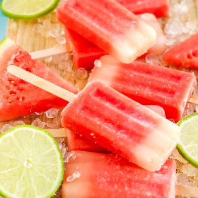 Close up photo of watermelon popsicles on a wooden cutting board with ice and lime slices scattered around them.