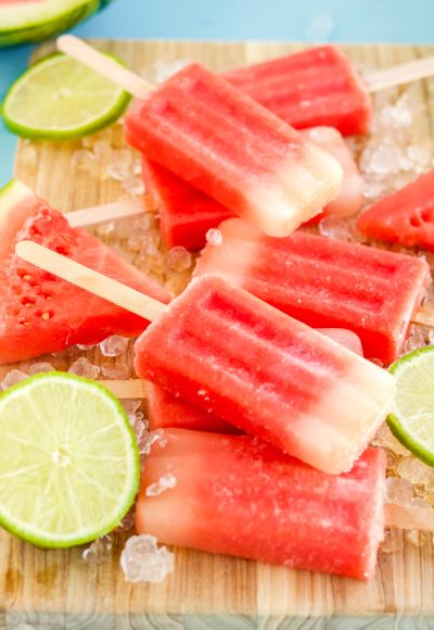 Close up photo of watermelon popsicles on a wooden cutting board with ice and lime slices scattered around them.