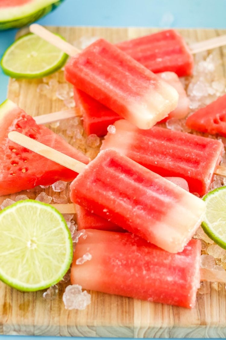 Close up photo of watermelon popsicles on a wooden cutting board with ice and lime slices scattered around them.