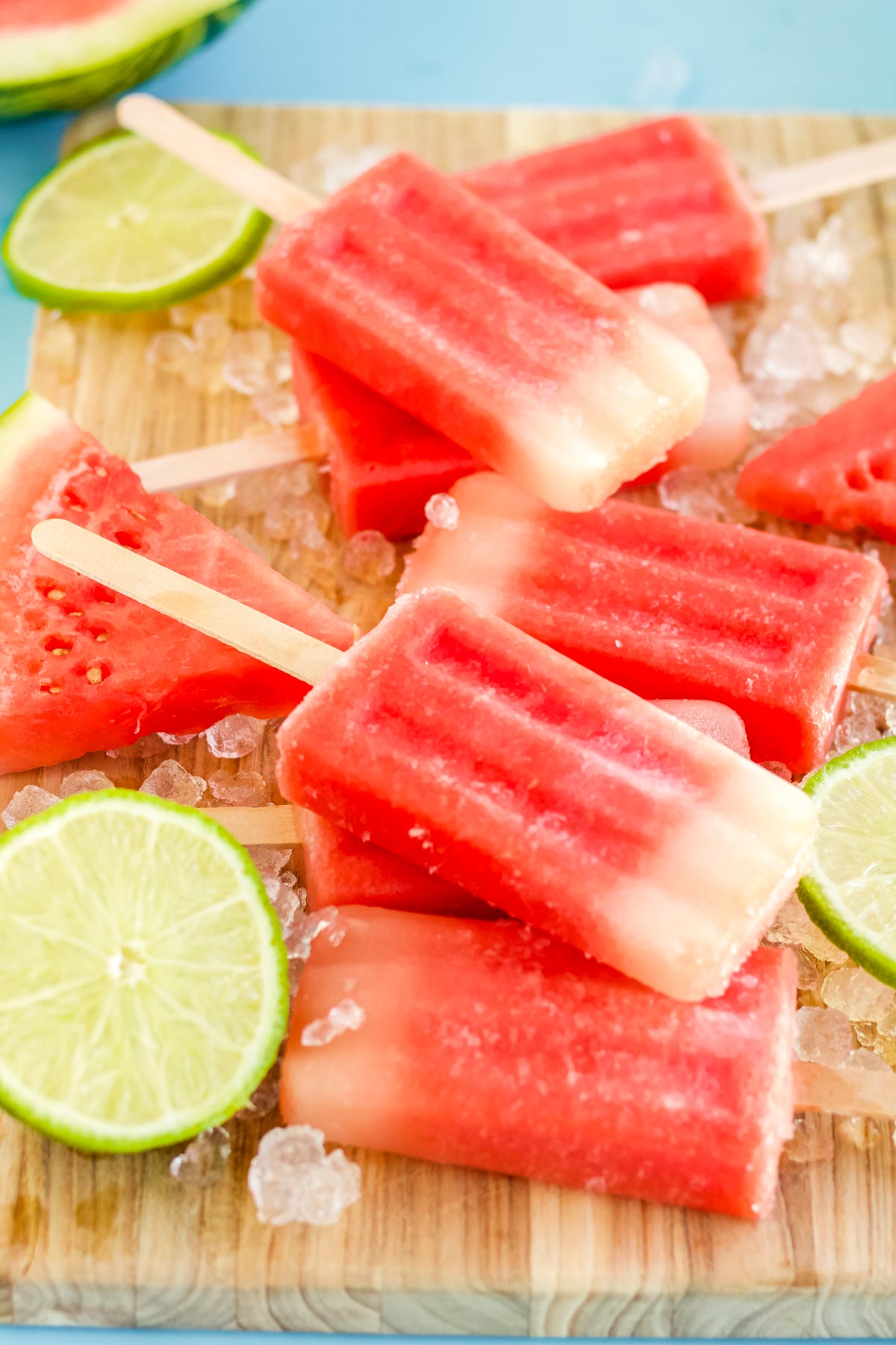 Close up photo of watermelon popsicles on a wooden cutting board with ice and lime slices scattered around them.