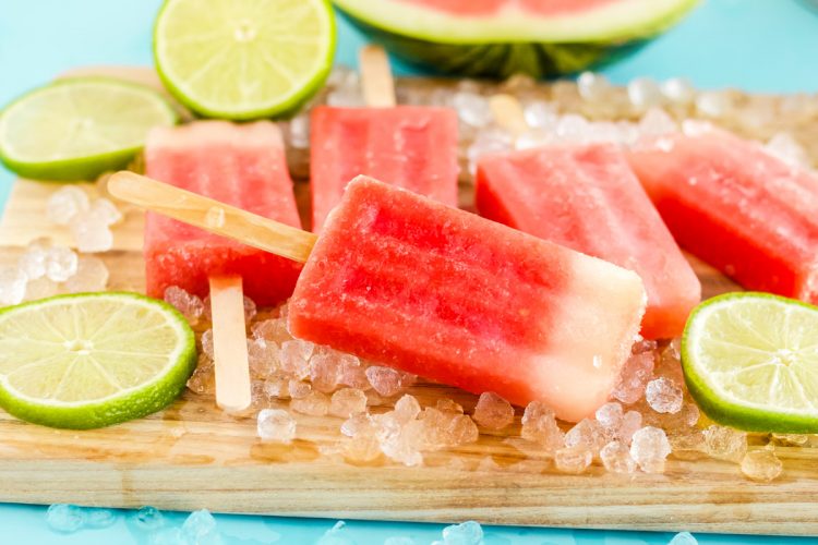 Watermelon popsicles on a wooden cutting board with lime slices and ice around them.