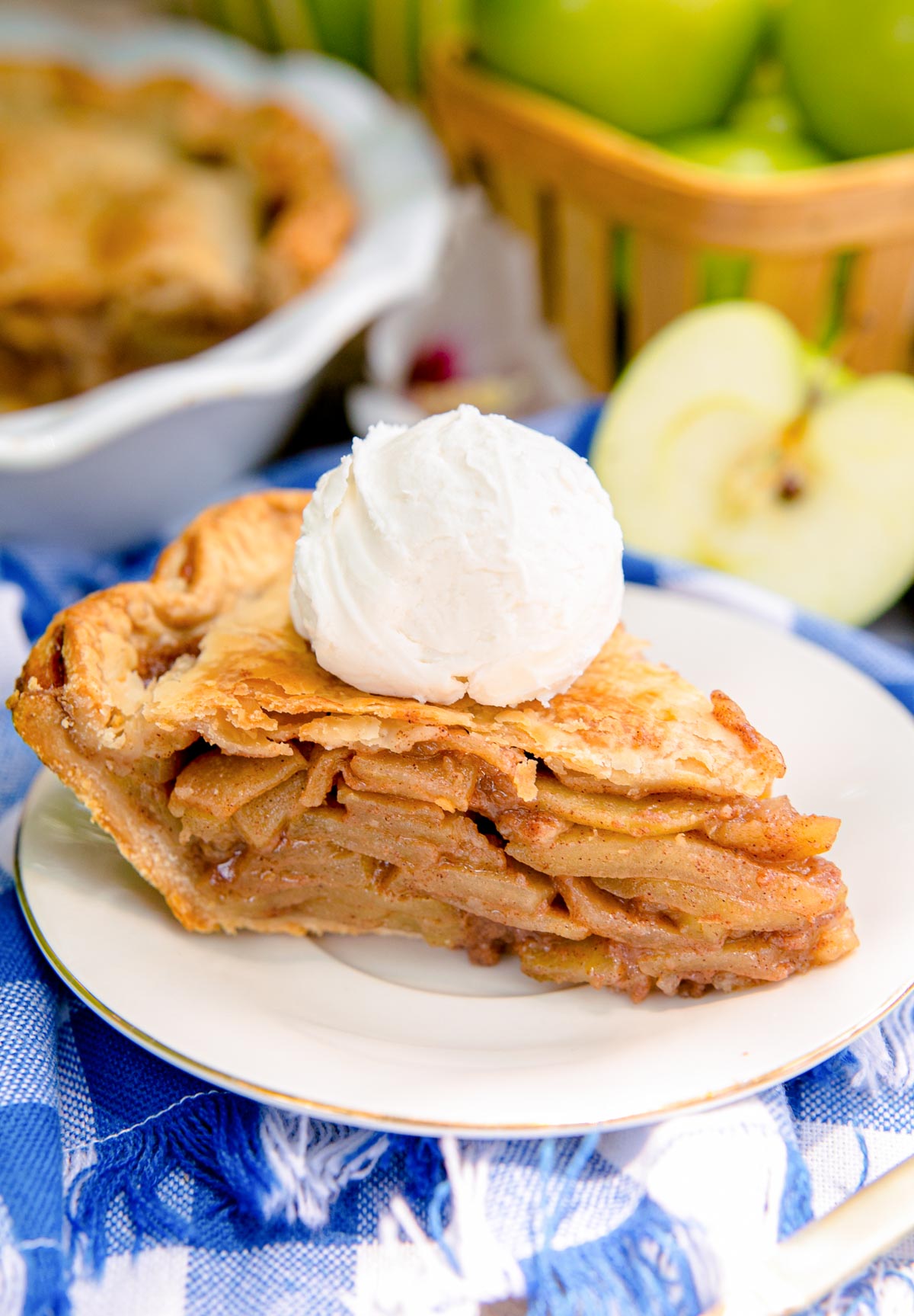 Close up photo of a slice of apple pie on a white plate on a blue gingham napkin with a basket of apples and the rest of the pie in the background.