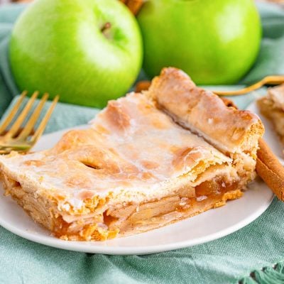 Close up photo of a piece of apple slab pie on a small white plate on a sage green napkin with cinnamon sticks and granny smith apples in the background.