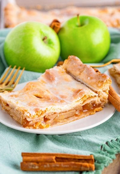 Close up photo of a piece of apple slab pie on a small white plate on a sage green napkin with cinnamon sticks and granny smith apples in the background.