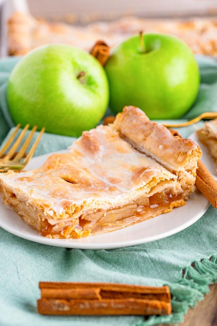 Close up photo of a piece of apple slab pie on a small white plate on a sage green napkin with cinnamon sticks and granny smith apples in the background.