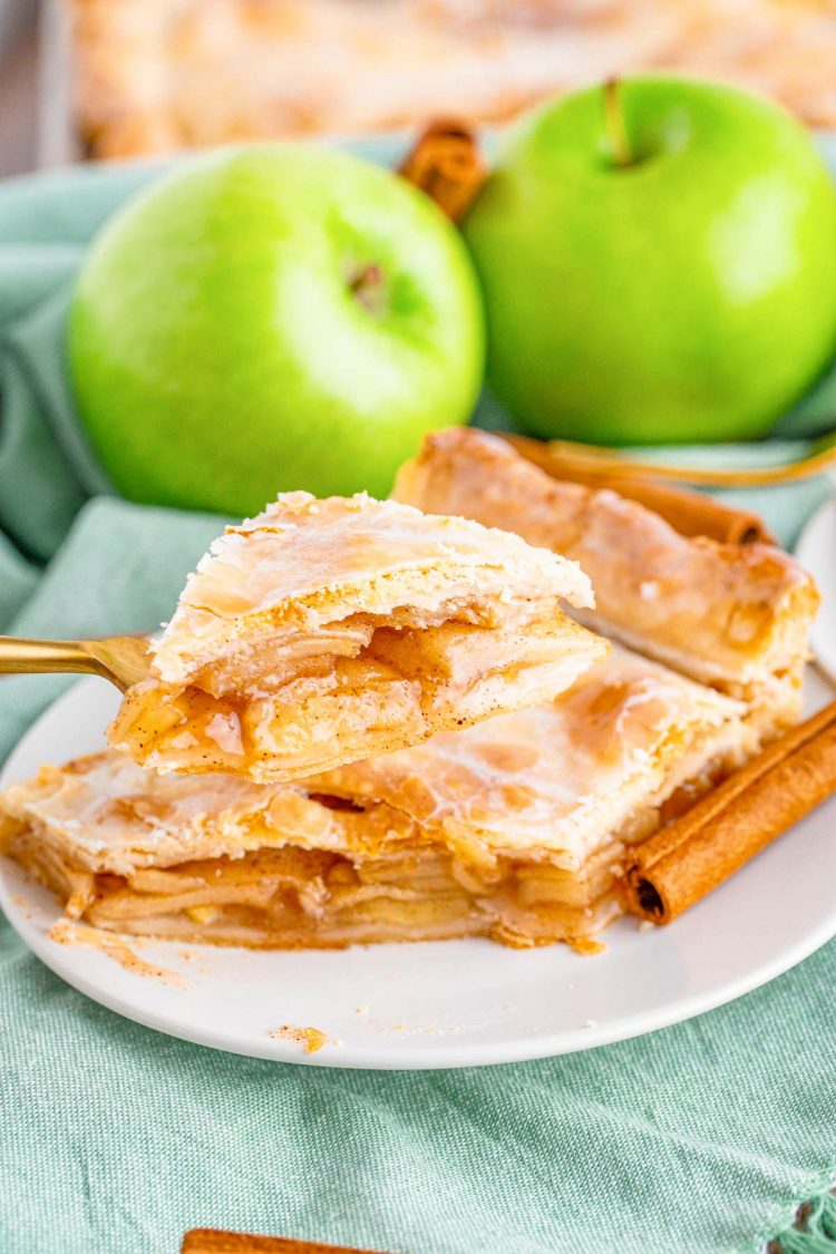 Close up photo of a slice of apple slab pie on a white plate on a blue napkin with a gold fork taking a bite from it. 