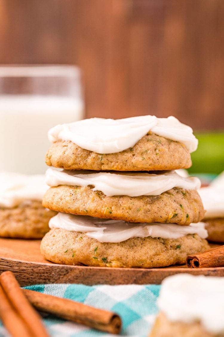 A stack of three frosted zucchini cookies on a wooden plate with more cookies and a glass of milk in the background.