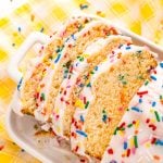 Close up photo of a slice of iced ice cream bread covered in rainbow sprinkles on a white plate on a yellow napkin.