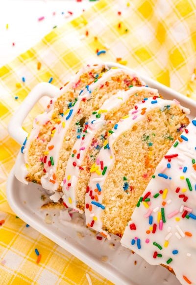 Close up photo of a slice of iced ice cream bread covered in rainbow sprinkles on a white plate on a yellow napkin.