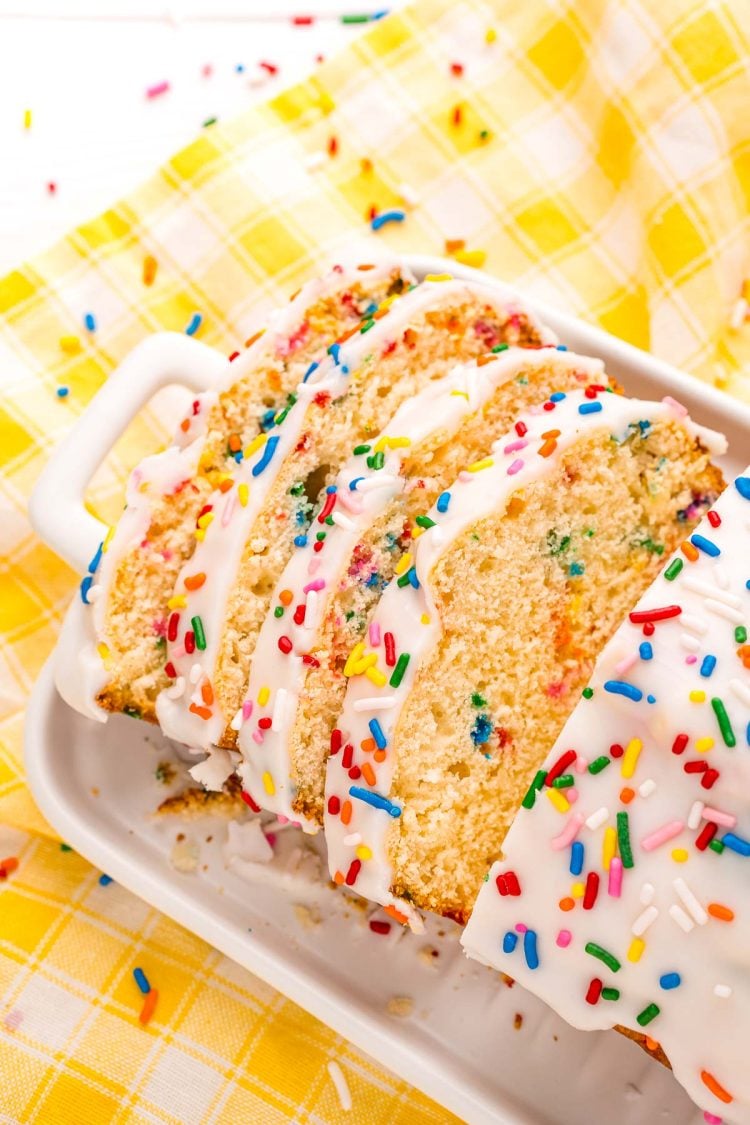 Close up photo of a slice of iced ice cream bread covered in rainbow sprinkles on a white plate on a yellow napkin.