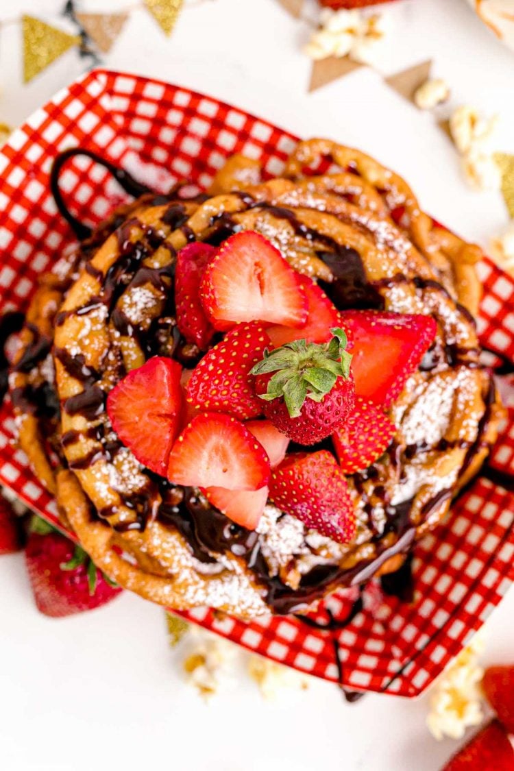 Overhead photo of funnel cakes in a red and white checkered food basket topped with strawberries and chocolate sauce.