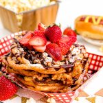 Close up photo of funnel cakes topped with powdered sugar, chocolate sauce, and fresh sliced strawberries in a paper tray.