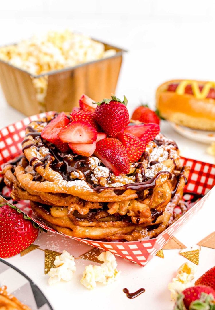 Close up photo of funnel cakes topped with powdered sugar, chocolate sauce, and fresh sliced strawberries in a paper tray.