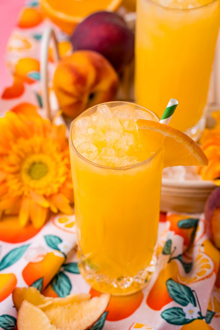 Close up photo of a fuzzy navel drink on a rattan tray with peaches and orange slices around it.