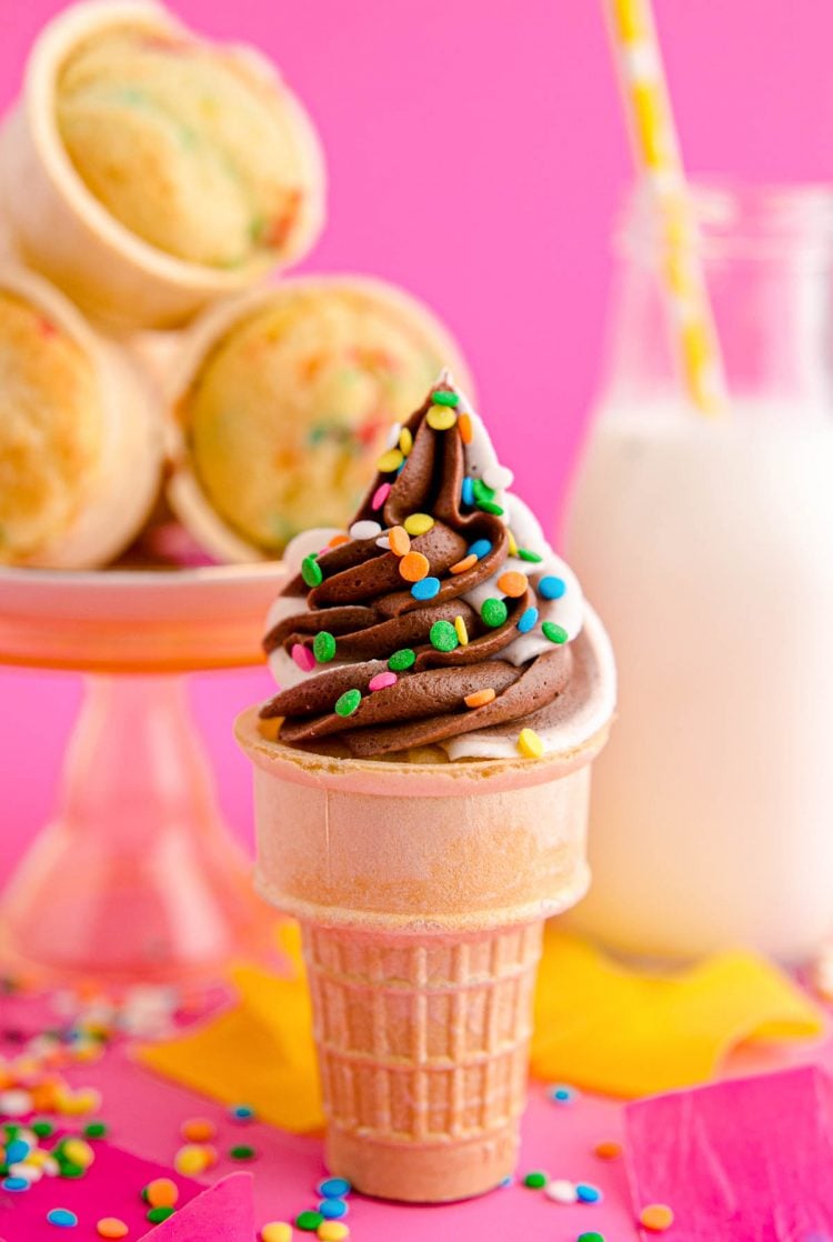 Close up photo of an ice cream cone cupcake topped with twist chocolate and vanilla frosting and colorful sprinkles. Bottle of milk and unfrosted cupcakes in the background. 