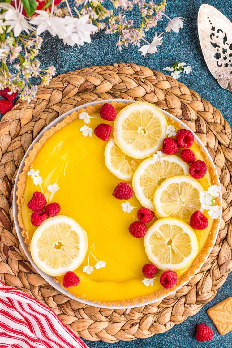 Overhead photo of a lemon tart on a wicker placemat with flowers and a red and white striped napkin around it.