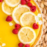 Overhead close up photo of a lemon tart on a wicker placemat topped with lemon slices, raspberries, and white flowers.