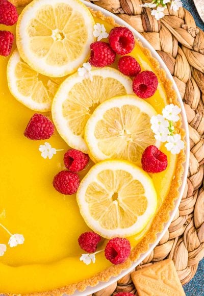 Overhead close up photo of a lemon tart on a wicker placemat topped with lemon slices, raspberries, and white flowers.