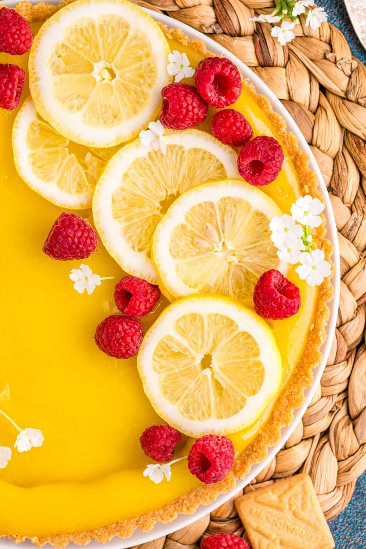 Overhead close up photo of a lemon tart on a wicker placemat topped with lemon slices, raspberries, and white flowers.