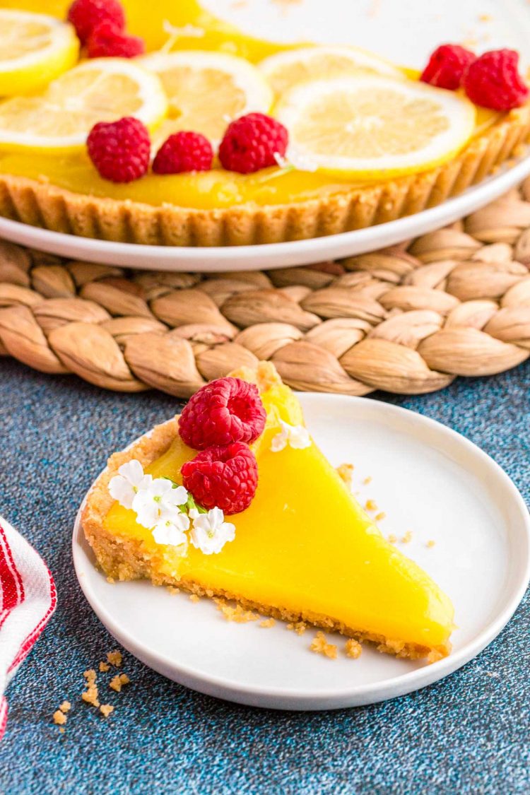 A slice of lemon tart garnished with raspberries on a white plate on a blue table with the rest of the tart on a wicker placemat in the background.