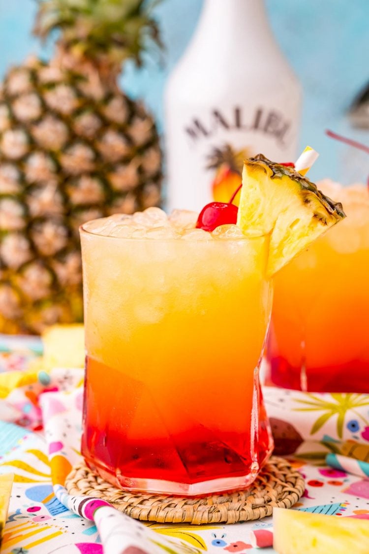 Close up photo of a Malibu sunset drink on a colorful napkin with another glass and pineapple in the background.