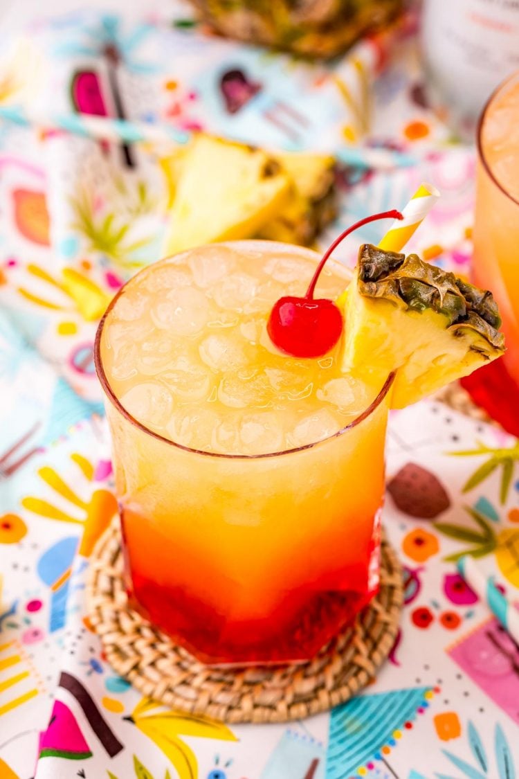Close up photo of a Malibu Sunset drink on a colorful printed napkin with pineapple wedges and another drink in the background.