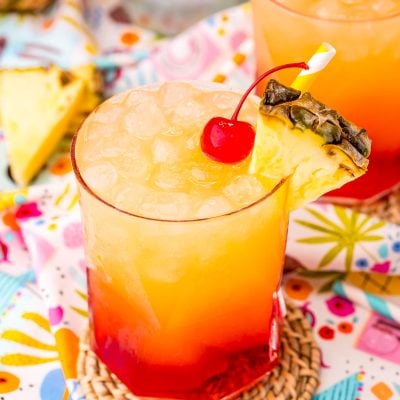 Close up photo of a Malibu Sunset drink on a colorful printed napkin with pineapple wedges and another drink in the background.