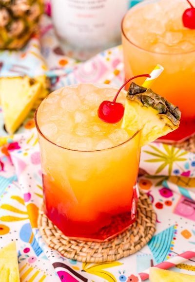 Close up photo of a Malibu Sunset drink on a colorful printed napkin with pineapple wedges and another drink in the background.