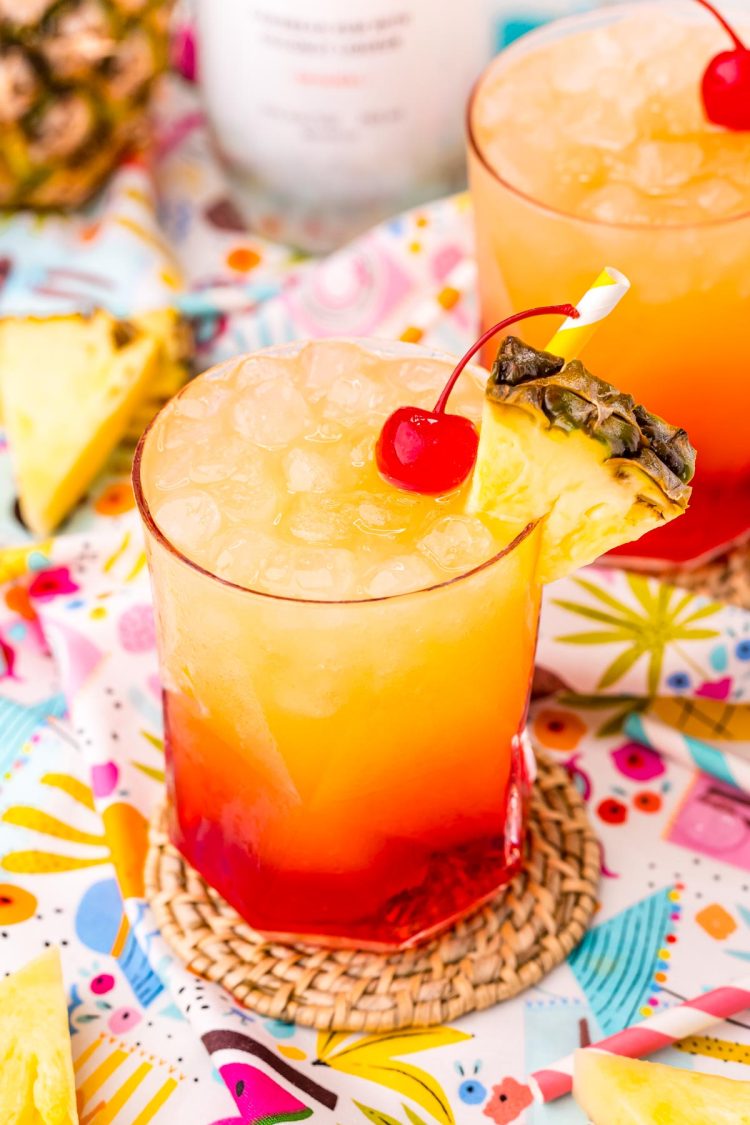 Close up photo of a Malibu Sunset drink on a colorful printed napkin with pineapple wedges and another drink in the background.