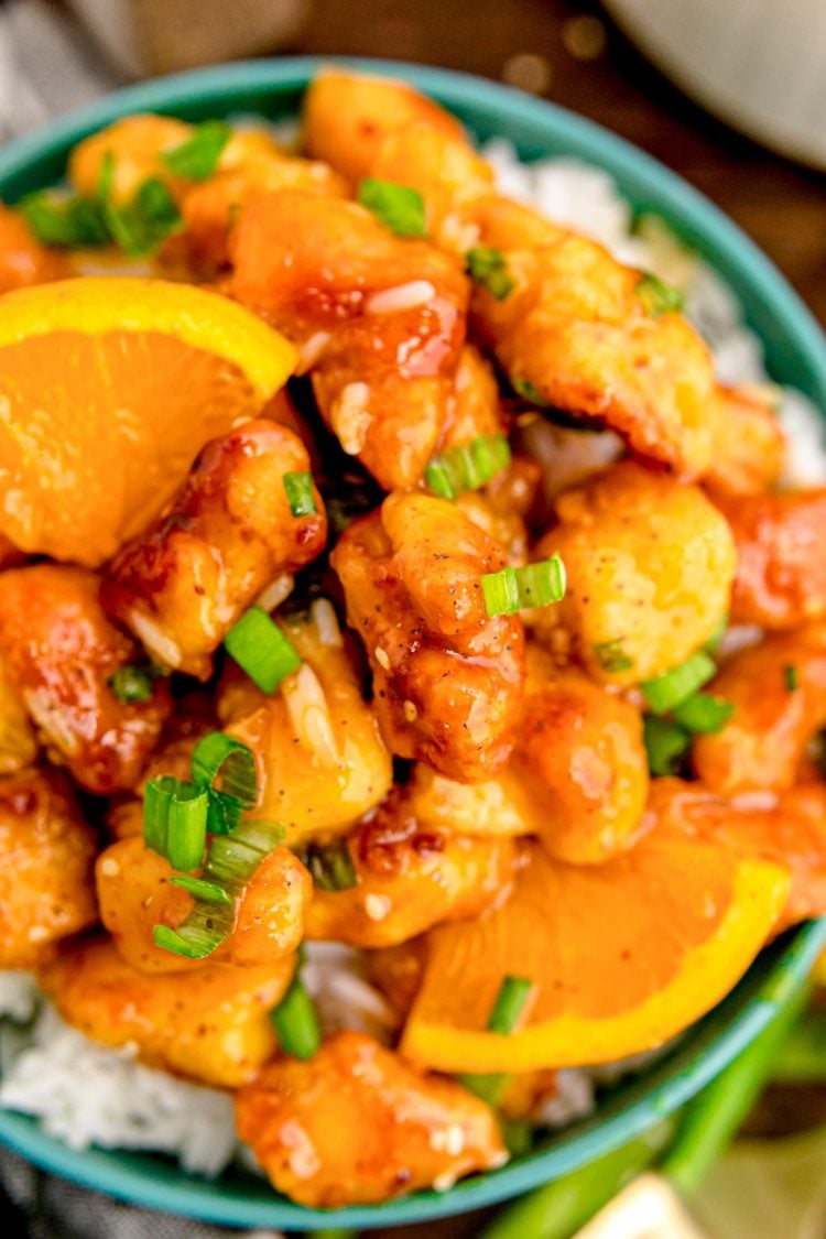 Close up photo of orange chicken on white rice in a teal bowl on a wooden table.