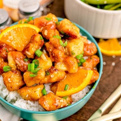 Close up photo of orange chicken on white rice in a teal bowl on a wooden table.