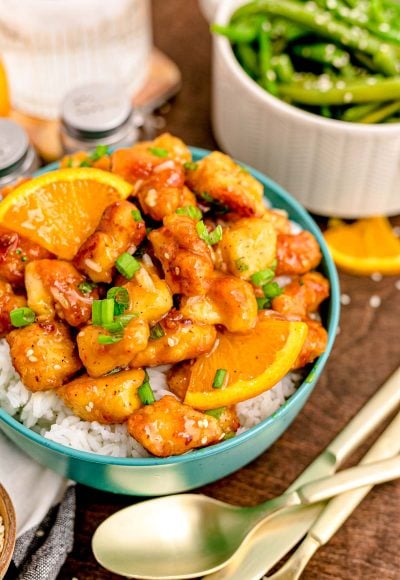Close up photo of orange chicken on white rice in a teal bowl on a wooden table.