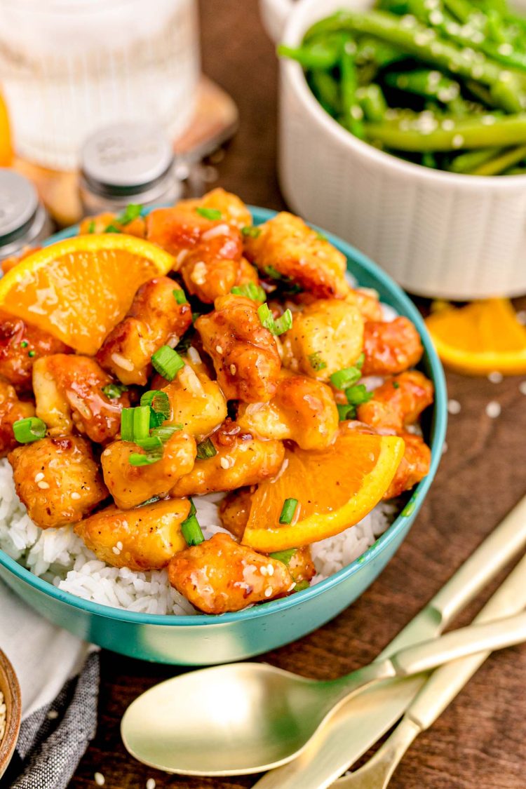 Close up photo of orange chicken on white rice in a teal bowl on a wooden table.