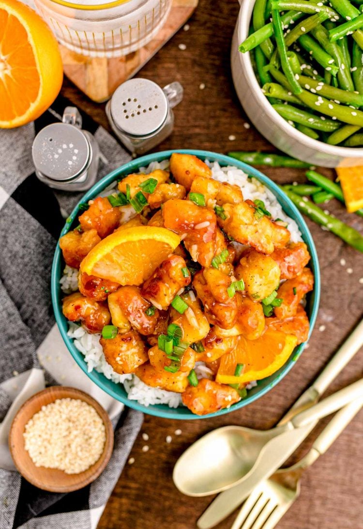 Overhead photo of a teal bowl filled with rice and orange chicken on a wooden table with utensils and ingredients around it.