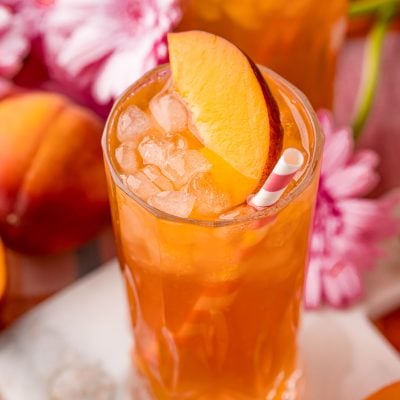 Close up photo of a glass of peach iced tea on a white marble coaster with peaches and pink flowers in the background.