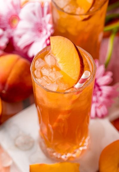 Close up photo of a glass of peach iced tea on a white marble coaster with peaches and pink flowers in the background.