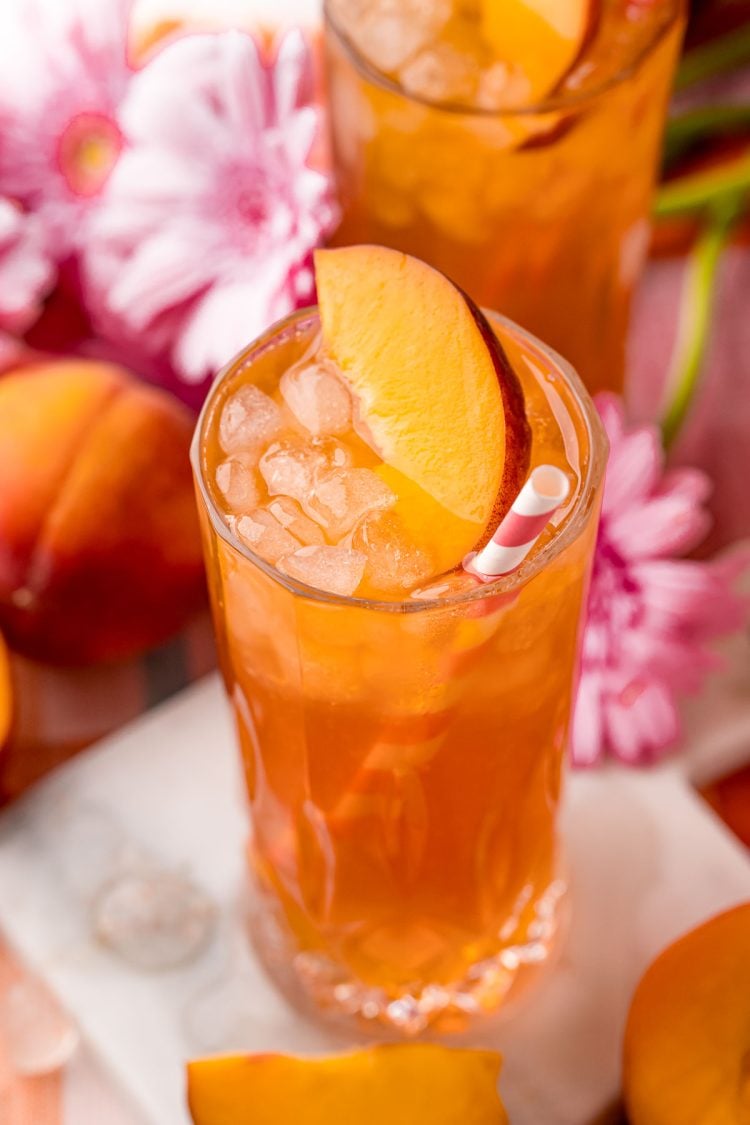 Close up photo of a glass of peach iced tea on a white marble coaster with peaches and pink flowers in the background.