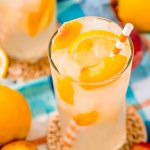 Close up photo of a glass with peach lemonade with lemons and peaches in it and an orange striped straw.