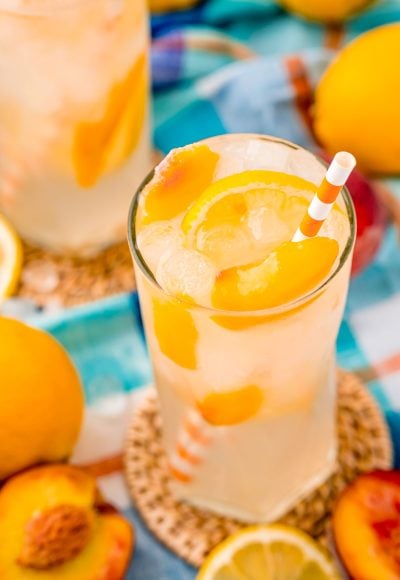 Close up photo of a glass with peach lemonade with lemons and peaches in it and an orange striped straw.