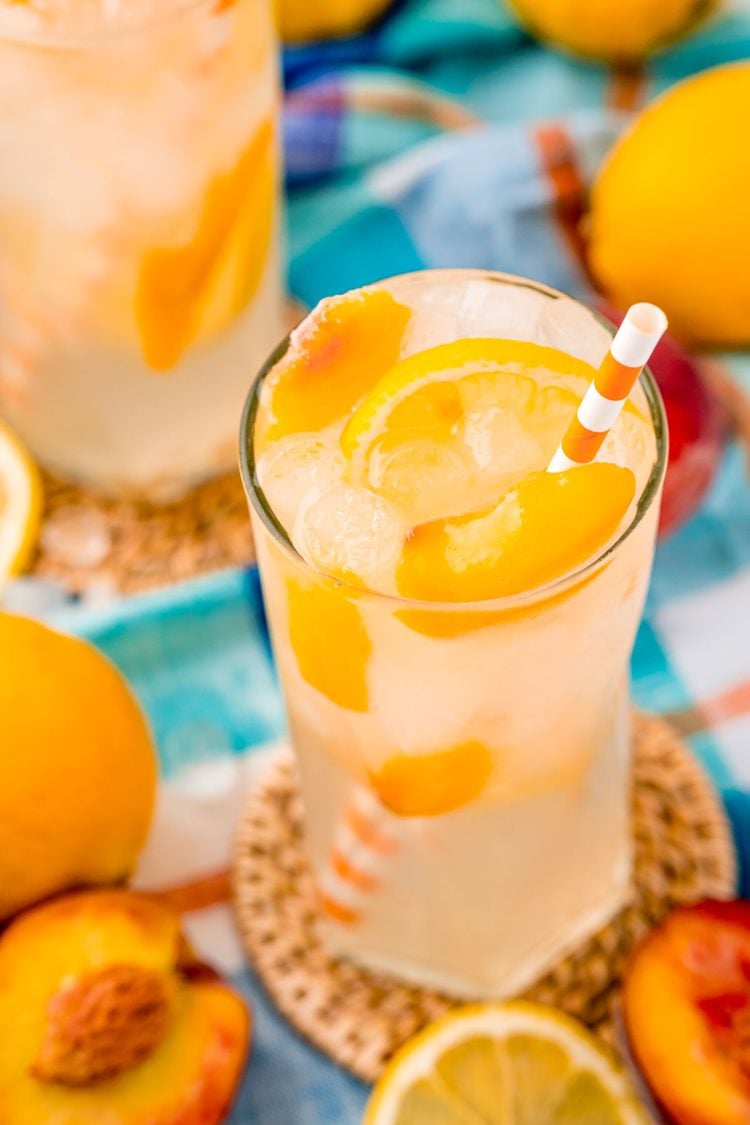 Close up photo of a glass with peach lemonade with lemons and peaches in it and an orange striped straw.