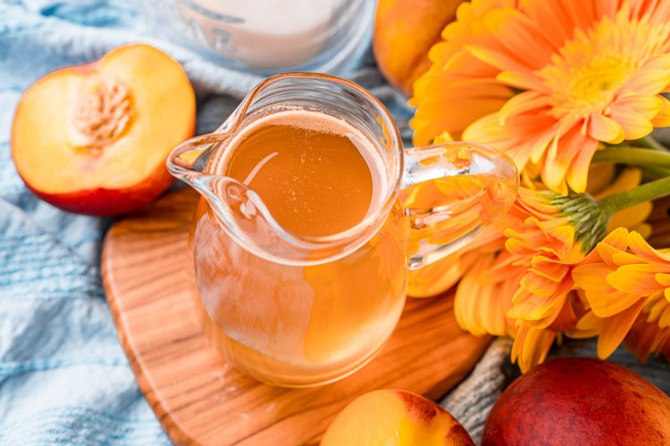 Horizontal photo of a small pitcher of peach simple syrup on a wooden cutting board.