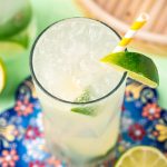 Close up photo of a ranch water cocktail on a colorful trivet with a white and yellow striped paper straw and lime wedges.