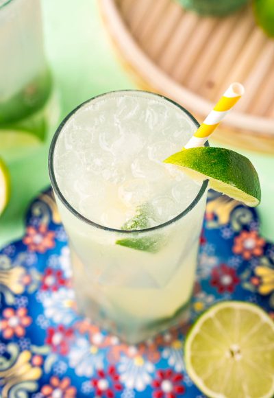 Close up photo of a ranch water cocktail on a colorful trivet with a white and yellow striped paper straw and lime wedges.