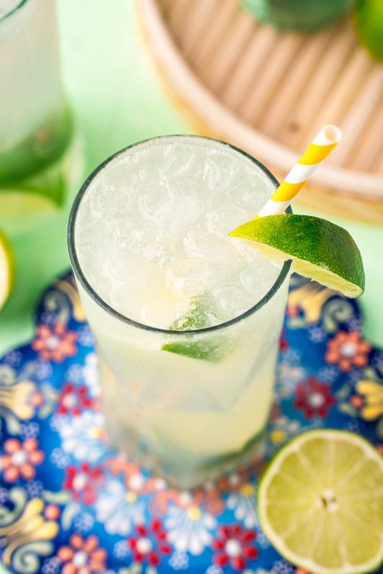 Close up photo of a ranch water cocktail on a colorful trivet with a white and yellow striped paper straw and lime wedges.