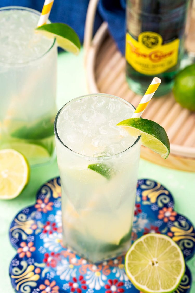 Highball glasses with ranch water in them on a colorful trivet surrounded by limes.