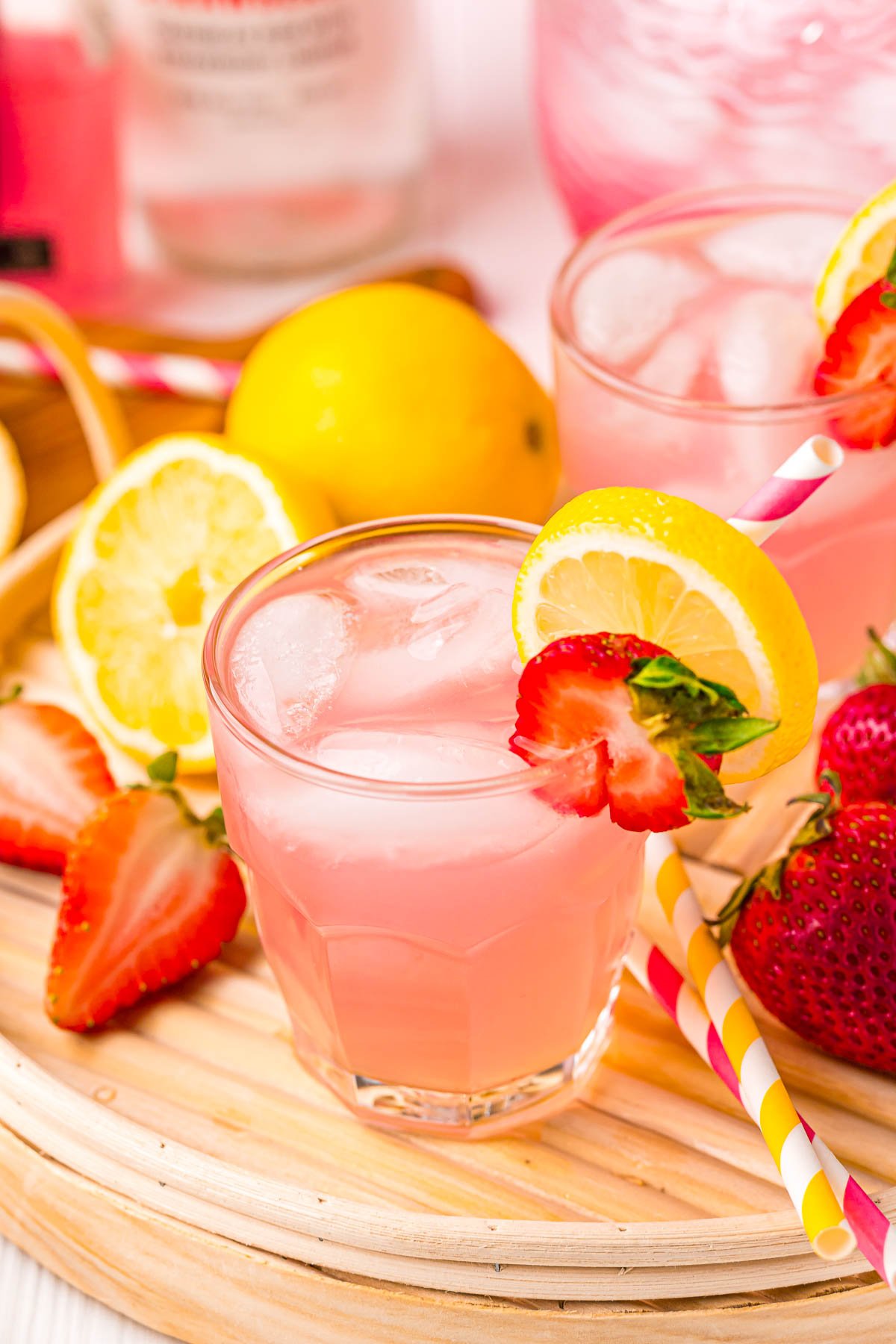 Close up photo of a glass of pink lemonade cocktail on a rattan tray garnished with lemon and strawberry.