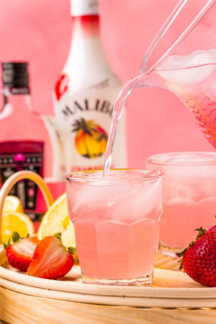 A pitcher pouring strawberry pink lemonade vodka into a short glass on a wicker tray with bottles of malibu and pink whitney in the background.